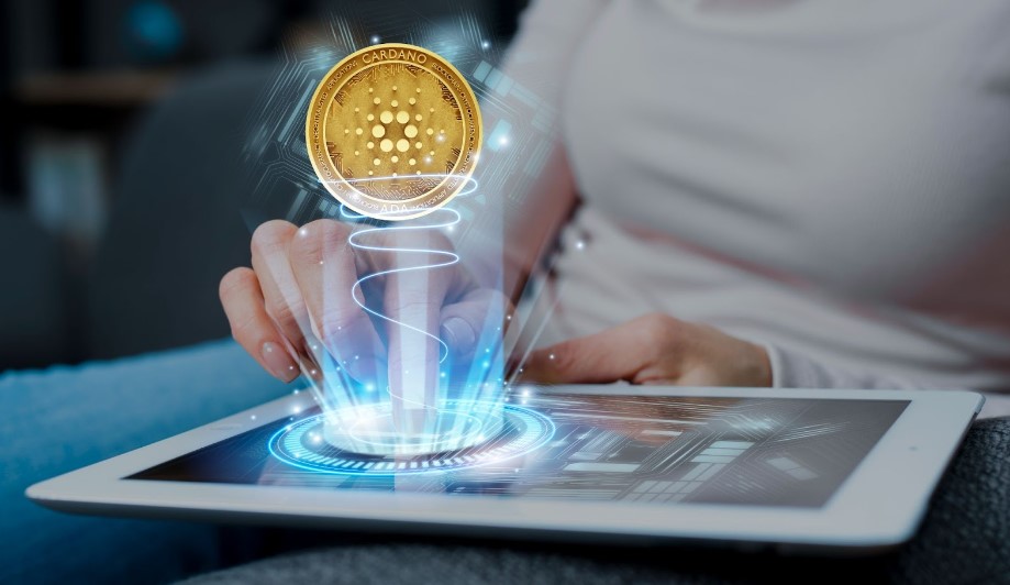 A person using a tablet from which a holographic projection of a Cardano cryptocurrency coin is emanating.
